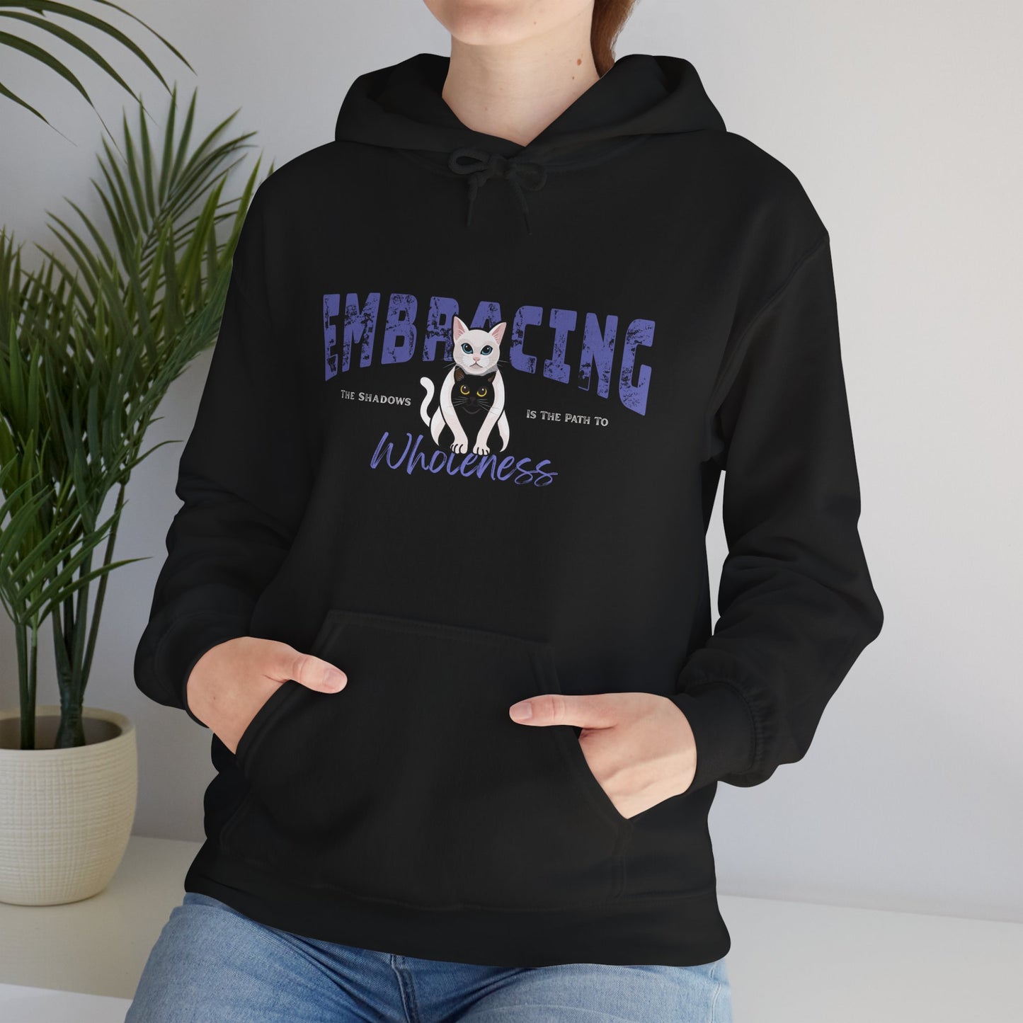 Person wearing a black hoodie with a graphic of white cat hugging black cat and text 'Embracing the shadows in the path to wholeness', standing indoors with a plant in the background.