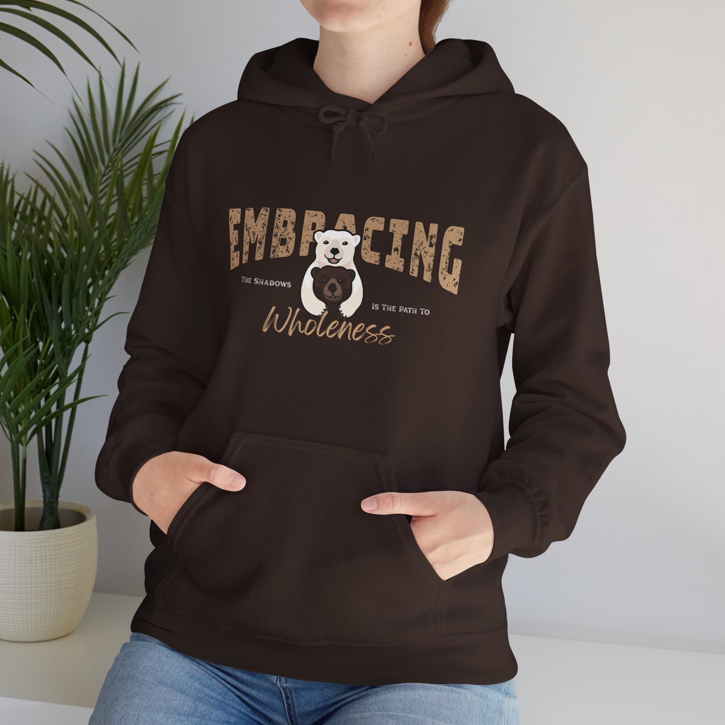 Person wearing a chocolate brown hoodie with a graphic of Polar bear hugging brown bear and text 'Embracing the shadows in the path to wholeness', standing indoors with a plant in the background.