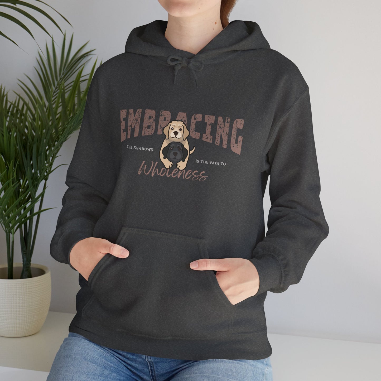 Person wearing a dark gray hoodie with a graphic of yellow Labrador Retriever hugging black Labrador Retriever and text 'Embracing the shadows in the path to wholeness', standing indoors with a plant in the background.
