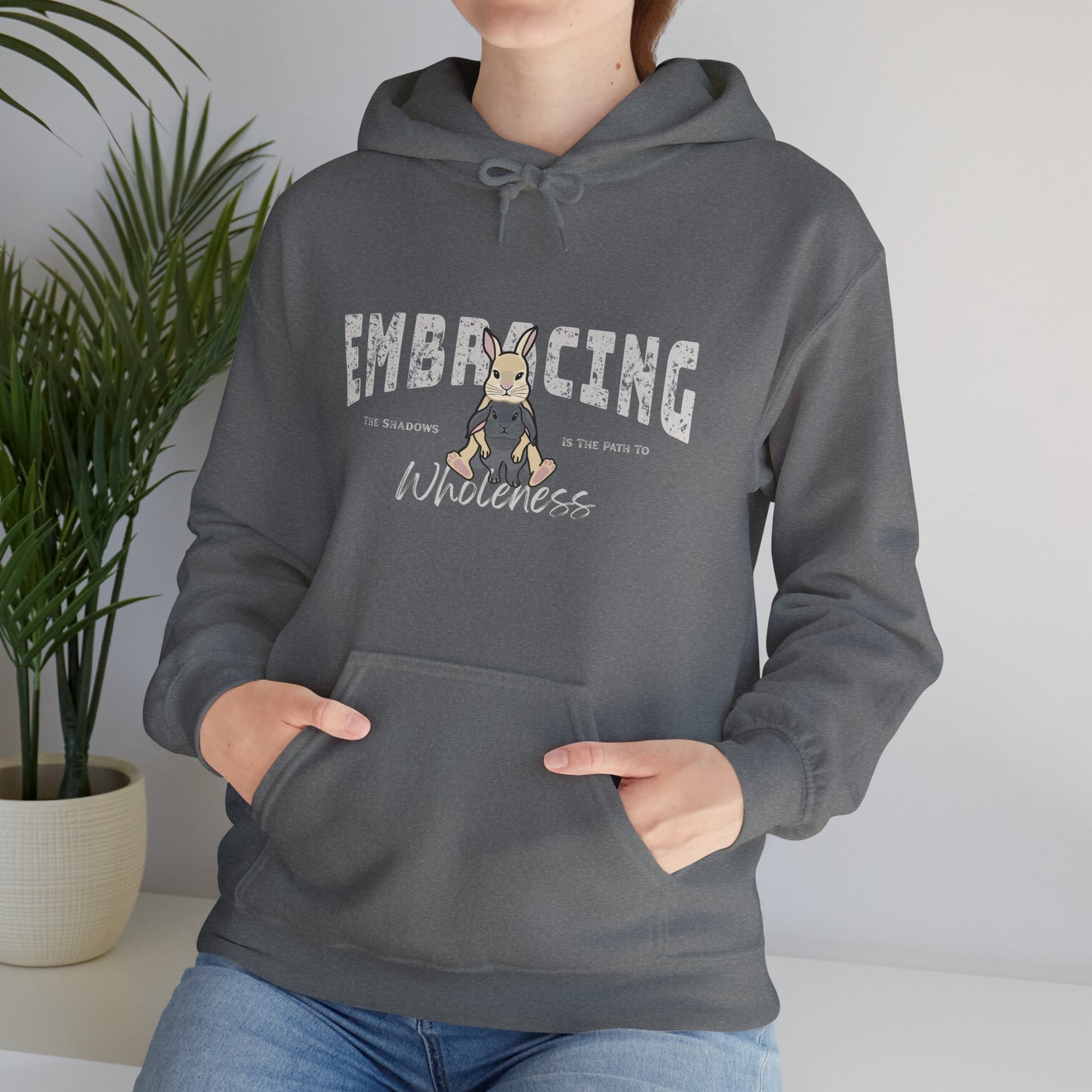 Person wearing a blue-gray hoodie with a graphic of cream Holland Lop rabbit hugging gray Holland Lop rabbit and text 'Embracing the shadows in the path to wholeness', standing indoors with a plant in the background.